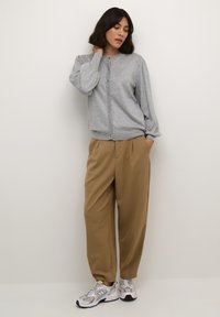 Cardigan gris à boutons sur le devant avec des manches bouffantes, associé à un pantalon plissé beige et des chaussures de sport grises avec des accents noirs.