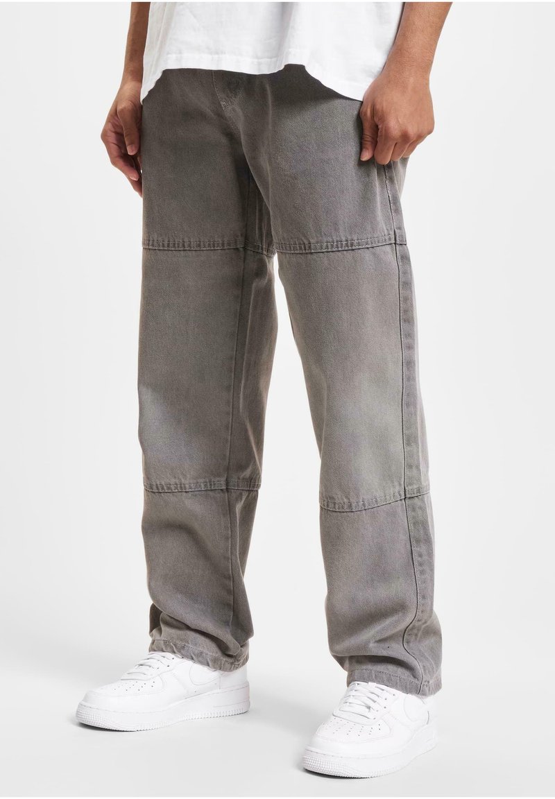 Jeune personne portant un pantalon patchwork gris ample et des baskets blanches, debout les mains détendues le long du corps.