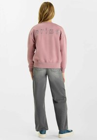 Roze sweatshirt met reliëflettering en een ribbelzoom, gecombineerd met loszittende grijze jeans en blauwe sneakers. Casual, relaxte look.