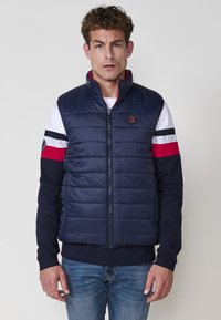 Chaleco acolchado azul marino con cuello alto, cierre de cremallera y costuras horizontales. Llevado sobre una camisa de manga larga de rayas rojas y blancas.