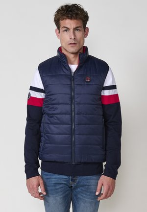Chaleco acolchado azul marino con cuello alto, cierre de cremallera y costuras horizontales. Llevado sobre una camisa de manga larga de rayas rojas y blancas.