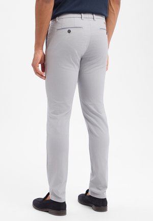 Mann trägt hellgraue, schmal geschnittene Hose und dunkle Loafers, steht mit locker an den Seiten hängenden Händen vor weißem Hintergrund.