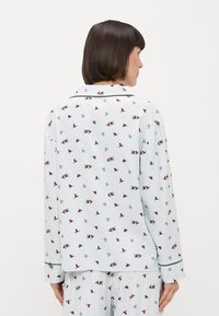 Svetle modrý pyžamový set s opakujícím se vzorem červených ptáků a zelených stromů. Měkká látka s límečkovým designem a kontrastním lemem.