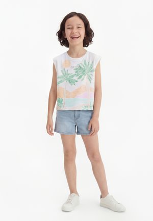 Uśmiechnięte dziecko z krótkimi kręconymi włosami, ubrane w biały bezrękawnik z tropikalnym nadrukiem, jasne szorty dżinsowe i białe trampki, stojące na białym tle.