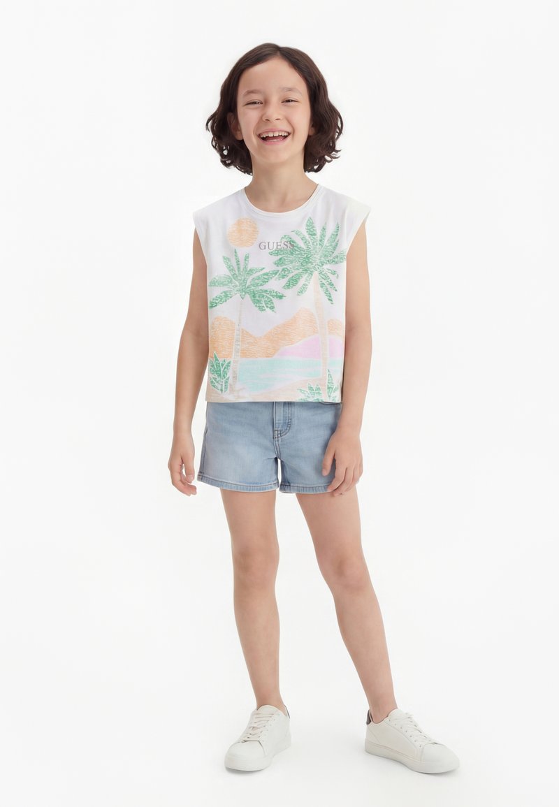 Χαμογελαστό παιδί με κοντά σγουρά μαλλιά, φορώντας λευκή αμάνικη μπλούζα με τροπικό σχέδιο, ανοιχτόχρωμα τζιν σορτς και λευκά αθλητικά παπούτσια, στέκεται σε λευκό φόντο.