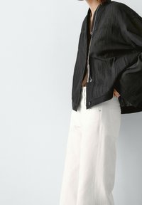 Black, textured jacket with a zip closure and wide sleeves, paired with high-waisted white trousers. Minimalistic design, no patterns.
