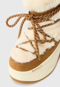 Beige platformlaars met een fluffy bovenkant, bruine suède accenten en getextureerde gouden hardware. Voorzien van gevlochten veters met witte details.