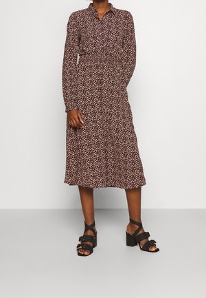 Kvinde iført en brun, blomsterprintet midi-kjole med lange ærmer og krave, kombineret med sorte sandaletter med hæl og anklenstropper.