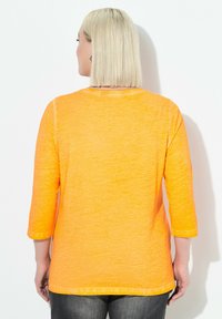 Donna con capelli biondi corti sta di fronte a una parete bianca, indossando una maglietta arancione a tre quarti di manica e pantaloni scuri.
