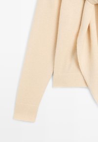 Blød beigefarvet sweater med ribkant ved kant og ærmer, lavet af et struktureret strikket stof, med et bundet detaljer foran.