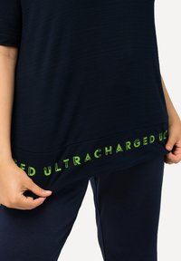 T-paita lyhythihaisena tummansinisenä, jossa on teksturoitu pinta. Vihreä "ULTRACHARGED" -teksti helman varrella. Mallin käsi nostaa kangasta hieman.