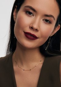 Złota biżuteria z delikatnym naszyjnikiem na łańcuszku, ozdobionym wielokolorowymi koralikami oraz pasującymi wiszącymi kolczykami, na neutralnym tle.