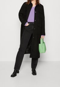 Abrigo de lana negro con dos bolsillos, usado sobre un suéter de canalé morado y jeans negros. Sosteniendo un bolso de cuero verde. Botas de tacón negras.