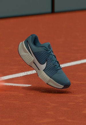 Zapatilla deportiva azul y blanca de Nike en el aire sobre una pista de tenis de arcilla con polvo rojo de arcilla volando cerca de una línea blanca de la pista.