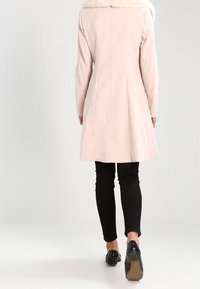 Ljusrosa kappa med en fuskpälskrage, figursydd design och utsvängd fåll. Bärs över svarta byxor och svarta skor. Slät textur, minimalistisk stil.