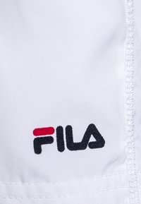 Logo de FILA bordado en hilo negro y rojo sobre tela blanca con costura visible a lo largo del borde derecho.