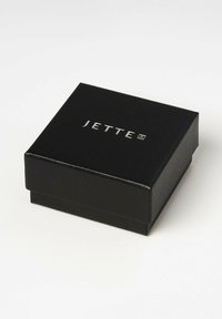 Schmuckkasten aus schwarzem Kunstleder mit glatter Textur, auf dem Deckel mit dem silbernen "JETTE"-Logo versehen. Quadratische Form mit einem Deckel, der mit Scharnieren befestigt ist.