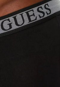 Un tissu noir avec une surface texturée présente une ceinture élastique grise contrastante affichant "GUESS" en lettres majuscules.
