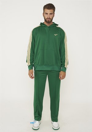 Uomo in piedi con tuta verde con strisce beige sulle maniche e scarpe da ginnastica bianche su sfondo bianco uniforme.