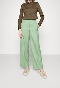 Camicia lunga marrone con colletto a punta, abbinata a pantaloni a vita alta e gamba larga verde menta. Sandali piattaforma beige con cinturini verdi.