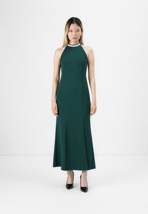 Woman wearing a sleeveless, dark green halter-neck gown with embellished collar, paired with black high heels, standing against white background.