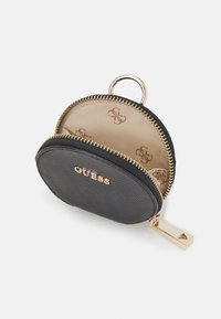Petit porte-monnaie rond en cuir noir avec fermeture éclair dorée, logo "GUESS" en or, intérieur beige avec motif de logos marron, et un anneau porte-clés doré.