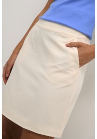 Nahaufnahme einer Person, die einen cremefarbenen Rock mit seitlichen Taschen trägt, eine Hand in einer Tasche, und ein blaues Oberteil vor einem neutralen Hintergrund.