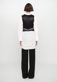 Persona con cabello corto y castaño, vistiendo una camisa blanca larga, un chaleco negro sin mangas y pantalones negros de pierna ancha, de espaldas.