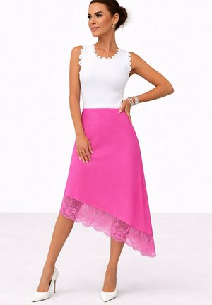 Femme portant un haut blanc sans manches à bords festonnés et une jupe rose asymétrique avec un ourlet en dentelle, posant avec la main sur la hanche et des talons blancs.