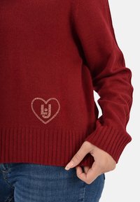 Maglione a maglia borgogna con orlo e polsini a coste, decorato con un design a forma di cuore in strass sulla parte anteriore.