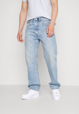 Person iført lyseblå løstsiddende jeans, hvide sneakers og en hvid skjorte, stående mod en ensfarvet lys baggrund.