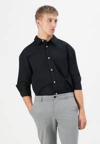 Jonge man met kort blond haar, gekleed in een zwart overhemd en lichtgrijze broek, met zijn handen in zijn zakken, kijkend opzij.