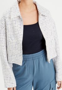 Geknipte jassie van textuurstof in een romige kleur met blauwe accenten. Heeft een ritssluiting, een brede kraag en losse mouwen. Gecombineerd met een zwarte top.