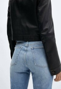 Chaqueta de cuero negra corta con una textura suave, combinada con vaqueros de tiro alto azul claro que tienen bolsillos traseros y detalles de costura sutiles.