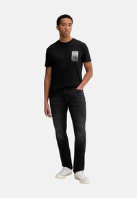 Camiseta de algodón negra con un estampado gráfico en el lado izquierdo del pecho, combinada con jeans oscuros y zapatillas blancas. Cuello redondo simple, corte holgado.