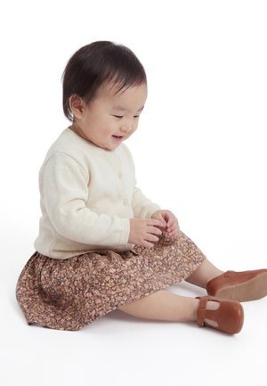 Toddler iført en cremefarvet sweater, brun blomster nederdel og brune sko, siddende på gulvet og smilende mens han kigger ned.