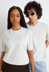 Ljusgrå t-shirts med korta ärmar, prydda med en rosa Adidas-logga. En modell bär leopardmönstrade byxor; den andra, denimshorts.