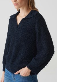 Pull over bleu marine en tissu texturé et duveteux, avec un col et un design en V. Coupe ample et longues manches, avec des poignets côtelés.