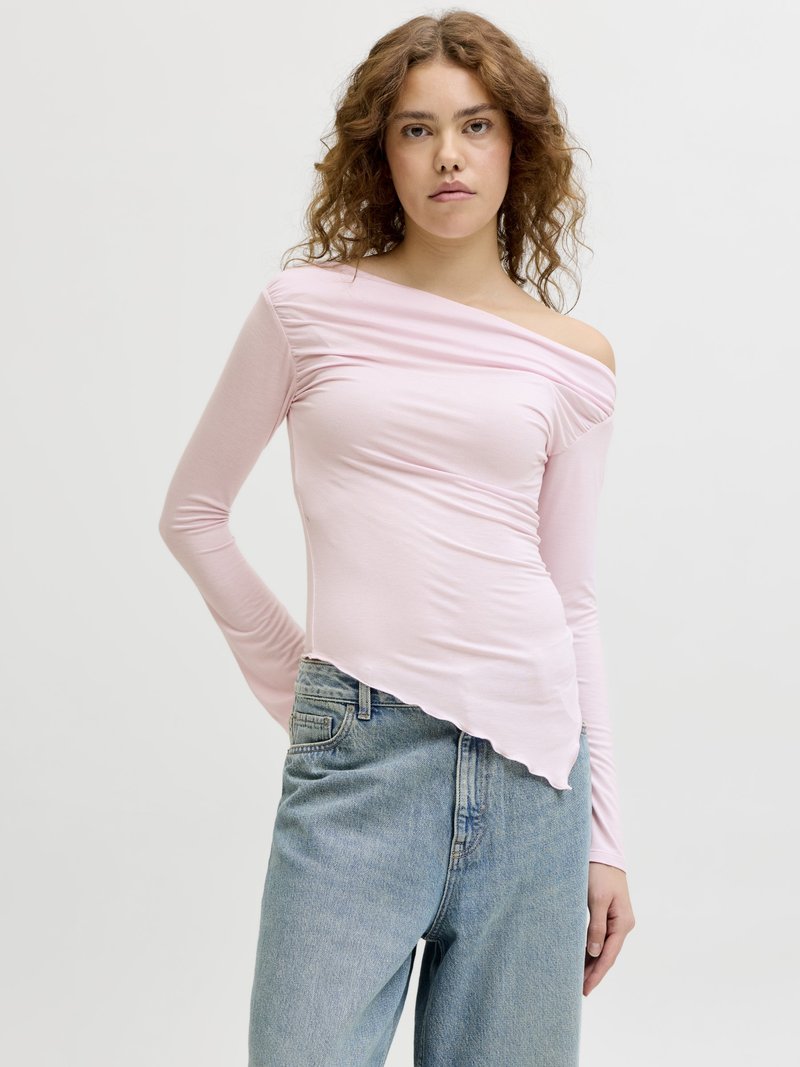 Νεαρή γυναίκα με κατσαρά μαλλιά που φοράει ανοιχτό ροζ πουκάμισο με μακριά μανίκια χωρίς ώμους και μπλε τζιν με ψηλή μέση, στέκεται μπροστά σε απλό φόντο.