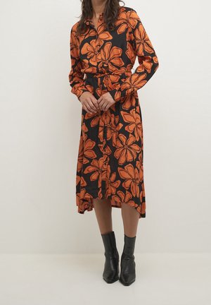 Femme portant une robe midi noire à grande impression florale orange, manches longues, devant boutonné, ceinture à la taille et bottines noires.