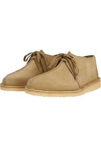 Beige mokasini nahkaa, joissa on pyöreä kärki, nauhakiinnitys edessä ja pehmeä kumipohja, sekä reunassa ommeltu yksityiskohta.