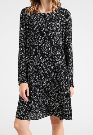 Frau trägt ein schwarzes langärmeliges Kleid mit einem weißen kleinen abstrakten Punktmuster, steht vor einem einfarbigen weißen Hintergrund.