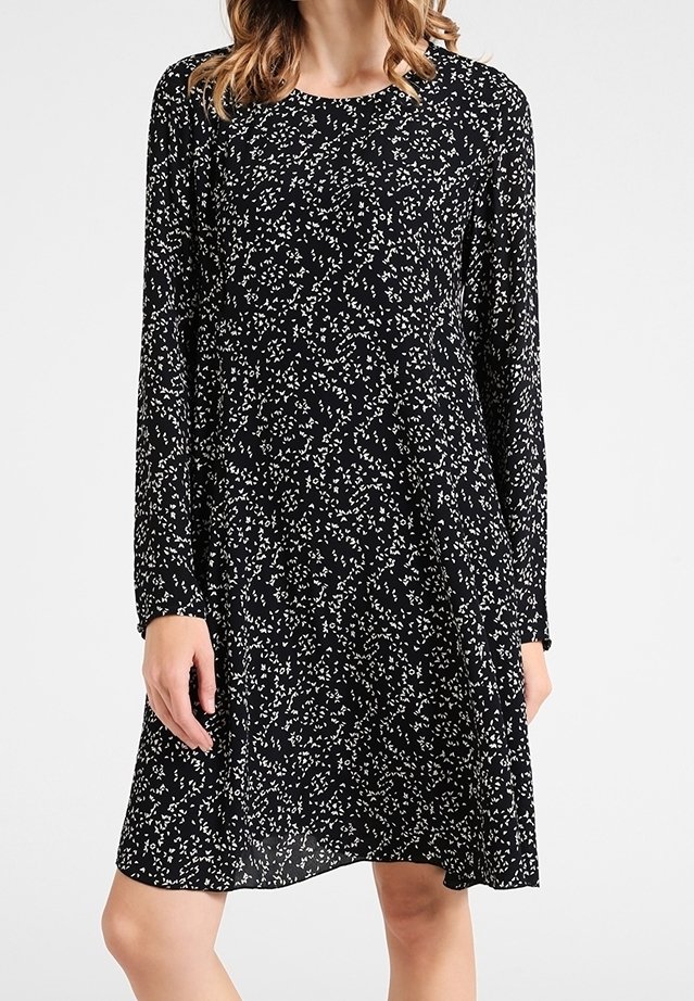 Femme portant une robe noire à manches longues avec un petit motif abstrait de points blancs, debout devant un fond blanc uni.