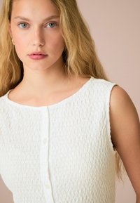 Haut sans manches blanc texturé avec un col rond et des boutons sur le devant. Tissu lisse avec un motif froissé subtil.