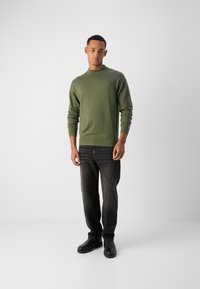 Pulover tricotat de culoare verde măsliniu, cu guler rotund, mâneci lungi și tiv striat, asortat cu blugi negri și pantofi negri. Design simplu, textură netedă.