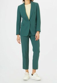 Ensemble deux pièces en teal avec une silhouette slim-fit, blazer à un bouton et pantalons fuselés, associés à des baskets blanches. Texture du tissu douce.