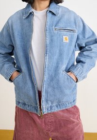 Lichtblauwe spijkerjack met een ritssluiting aan de voorkant, puntige kraag en borstzak. Draag het over een witte shirt, gecombineerd met gestructureerde kastanjebruine shorts.