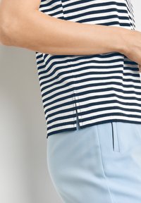 Námořnická a bílá pruhovaná bavlněná blůza s jemným bočním rozparkem je zkombinována se světle modrými kalhotami na míru, které se vyznačují hladkou strukturou a střihem.
