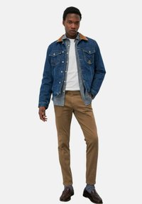 Giubbotto di jeans con colletto beige, camicia grigia sotto, maglietta bianca, pantaloni marroni e mocassini scuri; presenta due tasche sul petto.