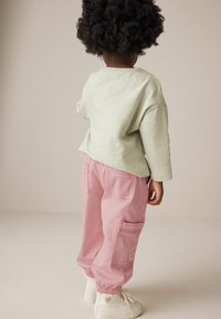 Cămașă lungă de culoare verde deschis, pantaloni cargo roz largi cu buzunare și adidași albi cu glezna înaltă, văzuți din spate.